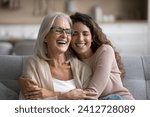 Happy excited senior mom and adult daughter woman having fun at home, hugging with heads touch, sitting on sofa, laughing with closed eyes, enjoying funny talk, leisure, family affection
