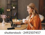 Side view young IT woman wearing orange shirt hold use laptop pc computer drink coffee sit alone at table in coffee shop cafe restaurant indoors work or study. Freelance mobile office business concept